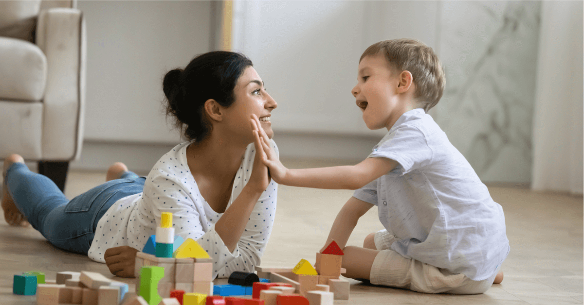 A young female nanny smiles and high-fives a happy little boy as they play together on the floor with colourful wooden blocks in a bright, cosy room